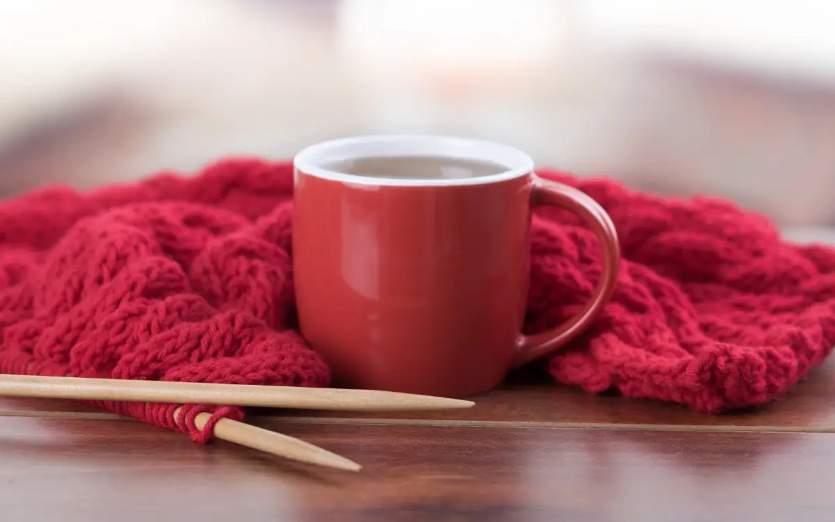 Eine rote Tasse mit Kaffee steht vor einem roten Strickstück mit hölzernen Nadeln auf einem Holztisch.